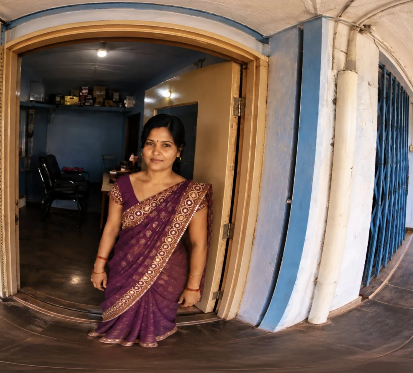 A shop owner in a purple saree stands in the lit doorway of her shop